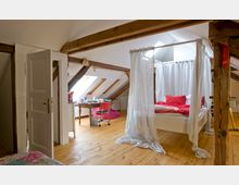 Dachgeschosszimmer mit sichtbaren Holzbalken, Holzdielenboden und Dachfenstern. Im Vordergrund steht ein Himmelbett mit weißen Vorhängen und roter Bettwäsche, daneben ein Schreibtisch mit Stuhl.