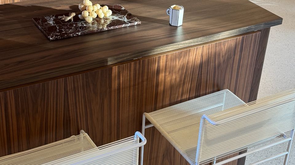 Modern kitchen island with dark wood paneling and a matching wood countertop, set on a light speckled floor. Two white wire-frame bar stools sit at the island, and a marble tray with grapes and a small cup rest on the surface, with wood cabinetry and a glass door in the background.