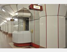 U-Bahn-Station mit futuristischer Architektur aus glatten weißen Paneelen und roten Akzenten, im Vordergrund ein kleiner geschlossener Kiosk mit beleuchtetem Schild.