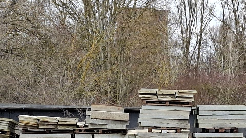 Gestapelte Betonplatten und Paletten auf einem Lagerplatz im Freien, davor eine dunkle Abdeckung oder Wand. Im Hintergrund stehen kahle Bäume und Sträucher, dahinter ragt ein großes, rechteckiges Gebäude teilweise verdeckt hervor.