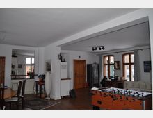 Wohnraum mit offenen Bereichen, weißen Wänden und Holzböden. Der Raum umfasst mehrere Fenstersätze mit Rundbögen, eine Küchenzeile, einen Esstisch, einen Kickertisch und einen Schrank aus dunklem Holz.