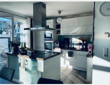 Moderne Küche mit weißen Hochglanz-Schränken, einem zentralen Kochfeld mit Insel-Dunstabzugshaube und großem Fenster mit Blick auf Häuser im Freien. Die Arbeitsflächen sind aus dunklem Holz, und links steht eine Obstschale auf der Kücheninsel.