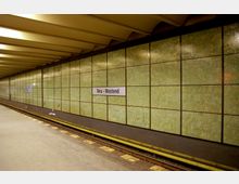 U-Bahn-Station "Neu-Westend" mit grünen Wandfliesen in rechteckigen Abschnitten, gelblich beleuchtetem Deckenpanel und einem Bahnsteig neben den Gleisen.