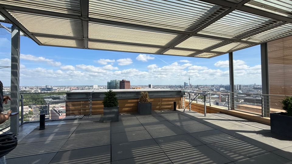 Rooftop terrace with large gray floor tiles, metal railings, and a slatted canopy that casts striped shadows across the deck. Planters line the edge, and the open side overlooks a wide city skyline under a partly cloudy sky.