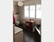 Badezimmer mit Tageslicht, ausgestattet mit einer Toilette, einem Waschbecken und einer Badewanne. Helle Fliesen an den Wänden, dunkler Fliesenboden und ein großes Fenster mit mattierter Verglasung; unter dem Fenster befindet sich ein Regal mit Körben und gefalteten Handtüchern.