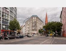 Straßenszene in einer Stadt mit einem Eckgebäude im Gründerzeitstil, das durch helle Fassaden und Balkone gekennzeichnet ist, im Hintergrund der rote Kirchturm einer Kirche. Am Straßenrand stehen geparkte Autos, und die Kreuzung ist von Ampeln und Zebrastreifen geprägt.