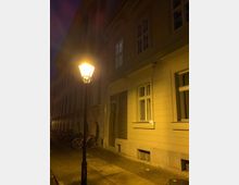 Eine straßenbeleuchtete Gasse bei Nacht mit einem historischen Gebäude auf der rechten Seite, charakterisiert durch weiße Fassade, helle Fensterrahmen und eine Holztür. Fahrräder sind entlang der Gebäudewand abgestellt, und eine Laterne spendet warmes Licht.