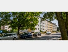 Mehrgeschossiges Wohngebäude in heller, farblich abgestufter Fassadengestaltung, umgeben von Bäumen. Im Vordergrund stehen parkende Autos entlang einer ruhigen Straße.