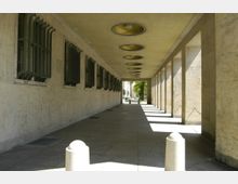 Überdachter Durchgang mit hellem Steinboden und -wänden, links gesicherte Fenster mit Metallgittern, rechts eine Reihe von rechteckigen Säulen. An der Decke befinden sich runde, goldene Einlassleuchten, und im Hintergrund sind Bäume und ein Gehweg sichtbar.
