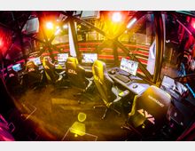 Professionelle E-Sport-Arena mit mehreren Schreibtischen, die jeweils mit Bildschirmen, Tastaturen, Headsets und Gaming-Stühlen ausgestattet sind. Der Raum ist in warmen Lichtern und futuristischem Design mit einem durchsichtigen Kuppelrahmen inszeniert.