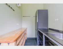 Schmaler Küchen- oder Hauswirtschaftsraum mit hellgrünen Wänden, grauen Fliesen und Edelstahl-Arbeitsfläche mit Spülbecken. An der Seite stehen ein hoher Kühlschrank und offene Metallregale, gegenüber eine lange Holzablage mit Untergestell sowie Wandhaken und Steckdosen.