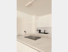 Moderne, minimalistische Küche mit weißen Schränken, einer großen Arbeitsplatte aus hellem Stein und einem integrierten schwarzen Kochfeld. Im Hintergrund eine schwarze Spüle, dekorative Elemente und eine schlichte Hängelampe an der Decke.