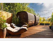 Holzterrasse mit einem kleinen, runden Holzsaunagebäude im Hintergrund; davor stehen zwei geschwungene Holzliegen mit gepolsterten Auflagen. Umgeben von Pflanzen und Lichterketten, in einem dicht bewachsenen Außenbereich.