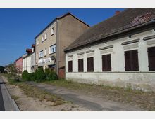 Straßenszene mit mehreren aneinandergereihten Gebäuden unterschiedlicher Fassadenstile: ein mehrstöckiges Wohnhaus mit einer Gaststättenbeschilderung und ein älteres Haus mit grüner Fassade und geschlossenen Holzläden. Im Vordergrund ein Gehweg mit Grasbewuchs und vereinzelt gepflanzten Bäumen, daneben eine Asphaltstraße.