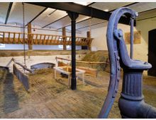 Innenraum eines traditionellen Viehstalls mit Strohboden und Futtertrögen, aufgeteilt durch Holzbalken und Metallketten. Im Vordergrund befindet sich eine alte Wasserpumpe, während im Hintergrund Heubündel und ein Gatter sichtbar sind.