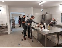 Ein moderner, heller Küchenraum mit weiß gefliesten Wänden und grauem Boden. Zwei Männer tragen Masken und arbeiten an einem hohen Edelstahltisch, der mit verschiedenen Flaschen, Schüsseln und Utensilien belegt ist; im Hintergrund sind weitere Arbeitsbereiche und Geräte wie ein Ofen sichtbar.