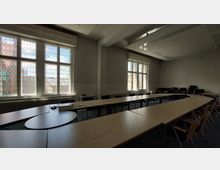 Großer Tagungsraum mit U-förmiger Tischanordnung und mehreren Stühlen. Die Fenster haben bunte, wabenförmige Scheiben, durch die Tageslicht einfällt, und an den Wänden sind Heizkörper sichtbar.