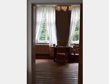 Ein Zimmer mit Holzfußboden, einem kleinen Tisch und zwei Holzstühlen vor einem großen Fenster mit weißen, lichtdurchlässigen Vorhängen. Im Hintergrund ist ein Heizkörper unter dem Fenster zu sehen, sowie ein Kronleuchter an der Decke.