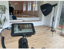 Heller Innenraum mit hellen Holzböden und großen Fenstern; im Hintergrund liegt eine erhöhte Sitz- oder Podestzone mit Tisch und Stehlampe. Im Vordergrund stehen Film-/Fotogeräte wie ein Kameramonitor auf Stativ und eine große Softbox-Leuchte neben einer Glastür zum Garten.