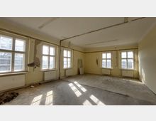 Leerer Raum mit gelb gestrichenen Wänden und mehreren großen Fenstern, die Tageslicht hereinlassen. Der Boden ist roh, die Wände zeigen Stellen mit Putzschäden, und Heizkörper befinden sich unter den Fenstern.