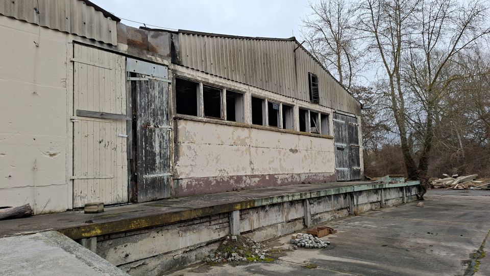 Verwittertes Industrie- oder Lagergebäude mit Wellblechdach, abblätternder Fassade, großen Holztoren und einer Reihe schmaler Fenster über einer erhöhten Laderampe. Davor liegt eine breite Betonfläche mit etwas Bauschutt; im Hintergrund stehen kahle Bäume.