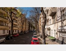 Straßenansicht einer Wohnstraße mit mehrstöckigen Gebäuden auf beiden Seiten, teils mit Balkonen. Entlang der gepflasterten Straße stehen Autos geparkt, und Bäume säumen den Gehweg, teilweise mit frühlingshaftem Laub.