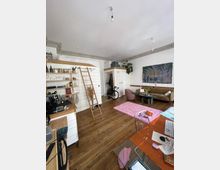 Ein Wohnzimmer mit Holzboden, minimalistischer Einrichtung und weiß gestrichenen Wänden. Zu sehen sind zwei eingebaute Holzschränke mit einer Leiter, ein Sofa mit einem rosa Teppich, ein Couchtisch, eine Küchecke mit Regalen sowie ein an die Wand gelehntes Fahrrad; die Decke ist einfach gehalten mit hängenden Lampen.