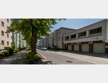 Eine ruhige Wohnstraße mit mehrstöckigen, pastellfarbenen Wohnhäusern auf der linken Seite und einer modernen Gebäudereihe mit Garagen im Erdgeschoss auf der rechten Seite. Ein gepflasterter Gehweg ist von Bäumen und kleinen Grünflächen gesäumt.