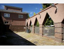 Backstein- und Putzgebäude mit symmetrischen Sprossenfenstern. Im Vordergrund ein gepflasterter Innenhof, begrenzt durch eine Mauer mit Spitzbögen und Metallgeländer; Lampen sind an der Mauer angebracht.