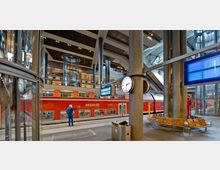 Ein moderner Bahnhof mit einer roten, doppelstöckigen Regionalzug der Deutschen Bahn am Bahnsteig. Im Vordergrund stehen Sitzbänke, eine runde Bahnhofsuhr und Anzeigetafeln, während im Hintergrund gläserne Aufzüge und Metallstützen die Architektur prägen.