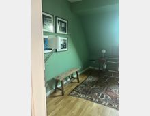 Ein kleiner Raum mit grünen Wänden, einem Holzboden und einem gemusterten Teppich. An der Wand hängen Schwarz-Weiß-Bilder, darunter eine Stehlampe und eine rustikale Holzbank; im Hintergrund ein Schreibtisch mit Stuhl und kleiner Lampe.