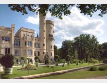 Historisches Schloss mit hellen Sandsteinfassaden, Zinnen und einem runden Turm, umgeben von einem gepflegten Garten mit Rasen, Blumenbeeten, Hecken und Topfpflanzen. Im Hintergrund stehen hohe Bäume unter einem blauen Himmel mit weißen Wolken.