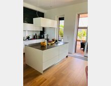 Moderne Küche mit weißer Kücheninsel und dunkler Arbeitsplatte, hängender rechteckiger Lampe darüber und dunkelgrüner Wandfliese im Hintergrund. Rechts führt eine Glastür zu einer Terrasse mit Holzboden und Pflanzen.