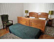 Schlafzimmer mit einem Doppelbett aus Holz im klassischen Stil, verziert mit Schnitzereien, flankiert von zwei passenden Nachttischen mit grünen Lampen. Links steht ein grüner Polstersessel, der Raum hat einen Holzboden, einen Teppich und gestreifte Tapeten.