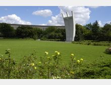 Eine Grünanlage mit einer großen, offenen Rasenfläche im Vordergrund, gesäumt von gelben Blumen und Sträuchern. Im Hintergrund ragt ein markantes Beton-Denkmal mit drei gebogenen Säulen empor, umgeben von Bäumen und einem mehrstöckigen Gebäude.