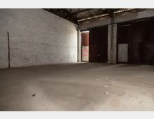 Leerer Innenraum einer Lagerhalle mit weiß verputzten Wänden, einem Betonboden und Metallträgern unter einem Wellblechdach. Eine teilweise geöffnete, große Metalltür lässt Licht herein, dahinter ist ein Schild mit der Aufschrift „Big Packs“ sichtbar.