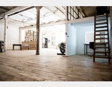 Loftartiges Interieur mit Holzboden, sichtbarer weißer Ziegelwand und Betonsäulen. Im Hintergrund befinden sich ein Bücherregal, rustikale Möbel und ein Motorrad, während rechts eine Holzleiter zu einer erhöhten Ebene führt und ein Schreibtisch an einer hellen Wand steht.