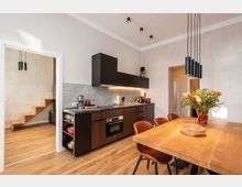 Moderne Wohnküche mit dunklen Hängeschränken und Holzfronten, grauer Arbeitsplatte und durchgehendem Fliesenspiegel sowie integrierten Geräten. Im Vordergrund steht ein großer Holztisch mit Stühlen unter schwarzen Pendelleuchten; links führt ein offener Durchgang zu einer Holztreppe.