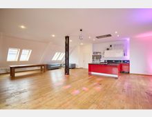 Modernes Loft mit offenem Raum, Holzboden und weiß gestrichenen Schrägwänden. Links ein langer Holztisch und Sofas vor mehreren Dachfenstern, rechts eine rote Küche mit weißen Hängeschränken, und in der Mitte eine schwarze Säule unter einer Discokugel.