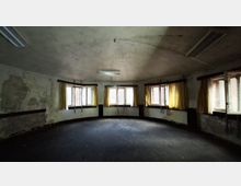 Leerer, runder Innenraum eines verlassen wirkenden Gebäudes mit abgenutzten Wänden, dunkelgrauem Boden und einer flachen Decke mit Leuchtstofflampen. Entlang der gebogenen Außenwand befinden sich mehrere Fenster mit teils gelben Vorhängen, durch die Tageslicht einfällt.