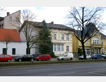 Straßenszene mit mehreren Wohnhäusern, darunter ein gelbes Gebäude mit dekorativem Giebel, ein weißes Haus mit großen Fenstern und ein kleineres Gebäude mit rotem Schrägdach. Vor den Häusern parken Autos entlang einer mehrspurigen Straße, wobei einige Bäume ohne Laub den Vordergrund säumen.