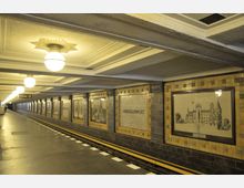 U-Bahn-Station mit Verkleidung aus cremefarbenen und braunen Wandfliesen, die Rahmen von historischen Zeichnungen enthalten, darunter der Schriftzug „Hohenzollernplatz“; helle Kugelleuchten hängen von der abgehängten Decke, und ein Bahnsteig mit gelber Sicherheitslinie erstreckt sich in die Tiefe.