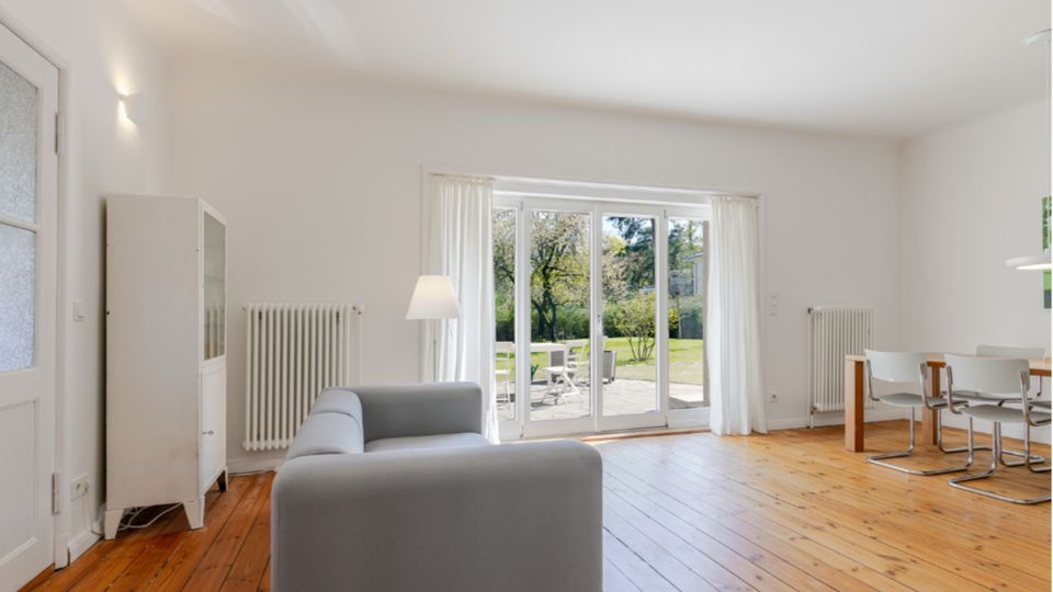 Heller Wohn- und Essbereich mit weiß gestrichenen Wänden und breiten Holzdielen, ausgestattet mit grauem Sofa, Vitrine und Esstisch mit Stühlen. Große verglaste Terrassentür mit weißen Vorhängen führt auf eine Terrasse und in einen grünen Garten.