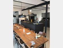 Moderne Wohnküche mit einer langen Holztafel und Stühlen in der Mitte des Raumes. Im Hintergrund befindet sich eine dunkle Küchenzeile mit eingebautem Backofen, Kochfeld, Dunstabzugshaube und Küchenschränken; die Decke zeigt sichtbare schwarze Tragstrukturen.