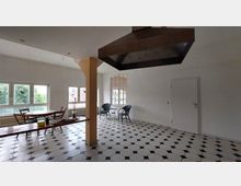 Geräumiger, heller Innenraum mit großen Fenstern, die viel Tageslicht hereinlassen, einem schwarz-weiß gefliesten Boden, einem zentralen Wandpfeiler und einer hängenden Dunstabzugshaube. Ein Holztisch mit Stühlen steht vor den Fenstern, im hinteren Bereich zwei Korbsessel mit kleinem Glastisch.