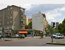 Straßenkreuzung in einer Stadt mit Wohn- und Geschäftsgebäuden, darunter ein Eckladen mit der Aufschrift „Fruchthaus“ und orangem Vordach. Im Hintergrund sind eine Kirche mit Uhrturm sowie weitere Häuser und Bäume zu sehen.