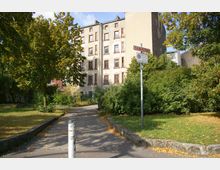 Ein kleiner Stadtpark mit gepflastertem Weg und Bäumen, im Hintergrund ein mehrstöckiges, älteres Wohngebäude mit hellen, verwitterten Fassaden und rot umrahmten Fenstern. Ein Verkehrsschild mit der Aufschrift „Feuerwehrzufahrt“ steht am Rand, umgeben von dichtem Grün.