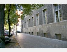 Straßenszene mit einem grauen, steinernen Gebäude mit großen Fenstern auf der rechten Seite, einem Gehweg aus Pflastersteinen und geparkten Autos entlang einer Baumreihe auf der linken Seite. Im Hintergrund sind einige Personen sowie weitere Gebäude zu sehen.