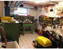 Ein gepflegter Werkstattraum mit Werkzeugwänden, auf denen verschiedene Handwerkzeuge wie Schraubenzieher, Zangen und Sägen organisiert hängen. Im Zentrum steht ein grüner Arbeitstisch mit einer Schraubzwinge, daneben befinden sich Reinigungsgeräte und Kabelrollen, während Rohre und Leitungen unter der Decke verlaufen.