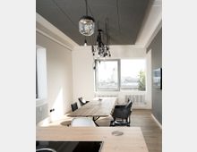 Modernes Esszimmer mit einem langen Holztisch und mehreren Stühlen, einer großen Fensterfront im Hintergrund, die auf eine städtische Umgebung blickt. Vom schwarzen Deckenpaneel hängen mehrere lose verkabelte Lampen herab, das Zimmer hat einen Holzboden und eine minimalistische Einrichtung.